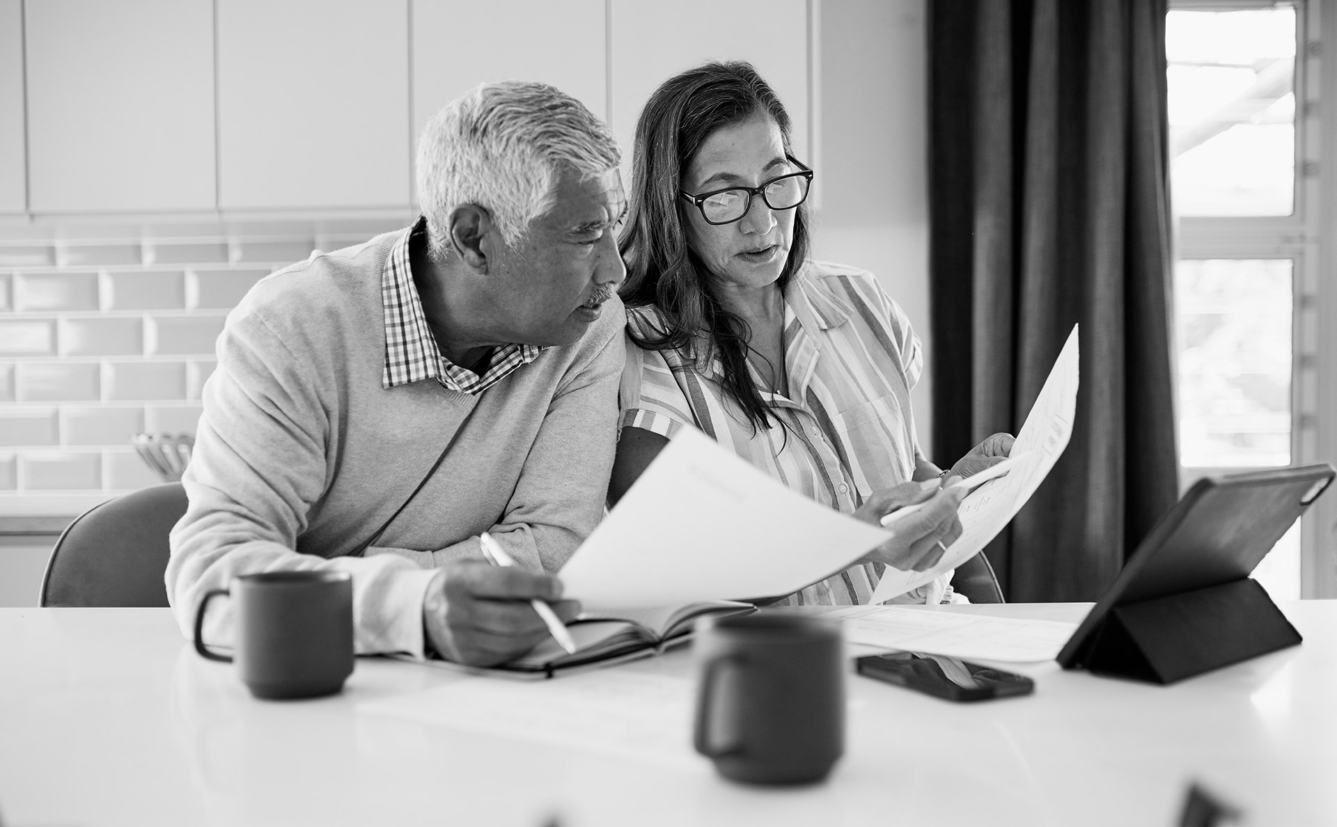 Older couple reviewing financial documents at a table with mugs and a tablet in front of them.