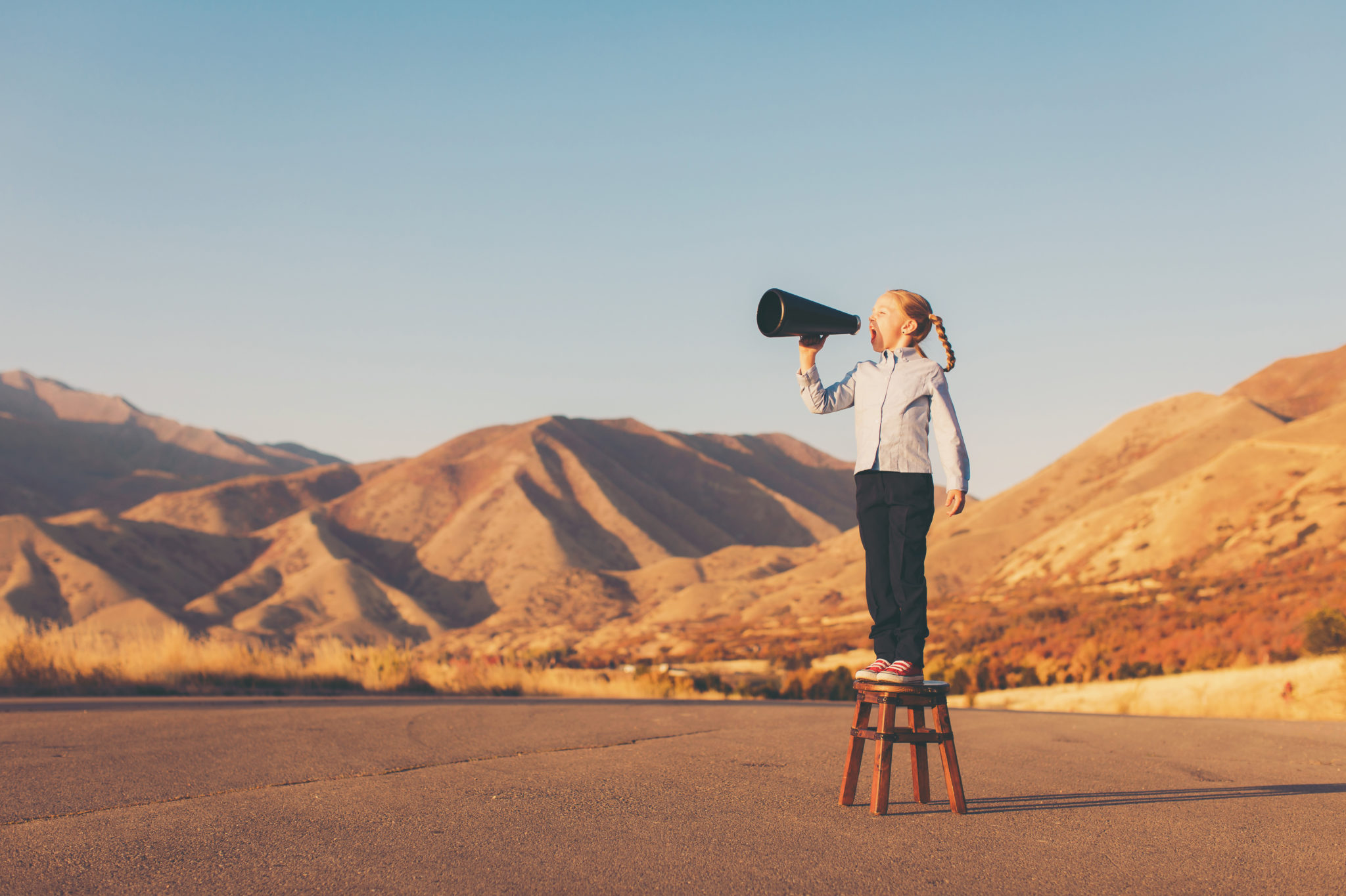 Girl standing on a stool using a megaphone in a scenic outdoor setting.