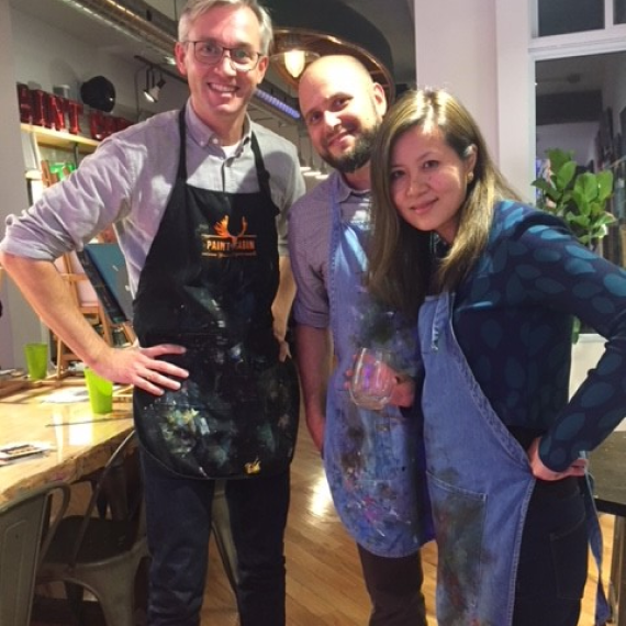 Three Ext. employees smiling together in a painting class, wearing aprons and standing in a studio.